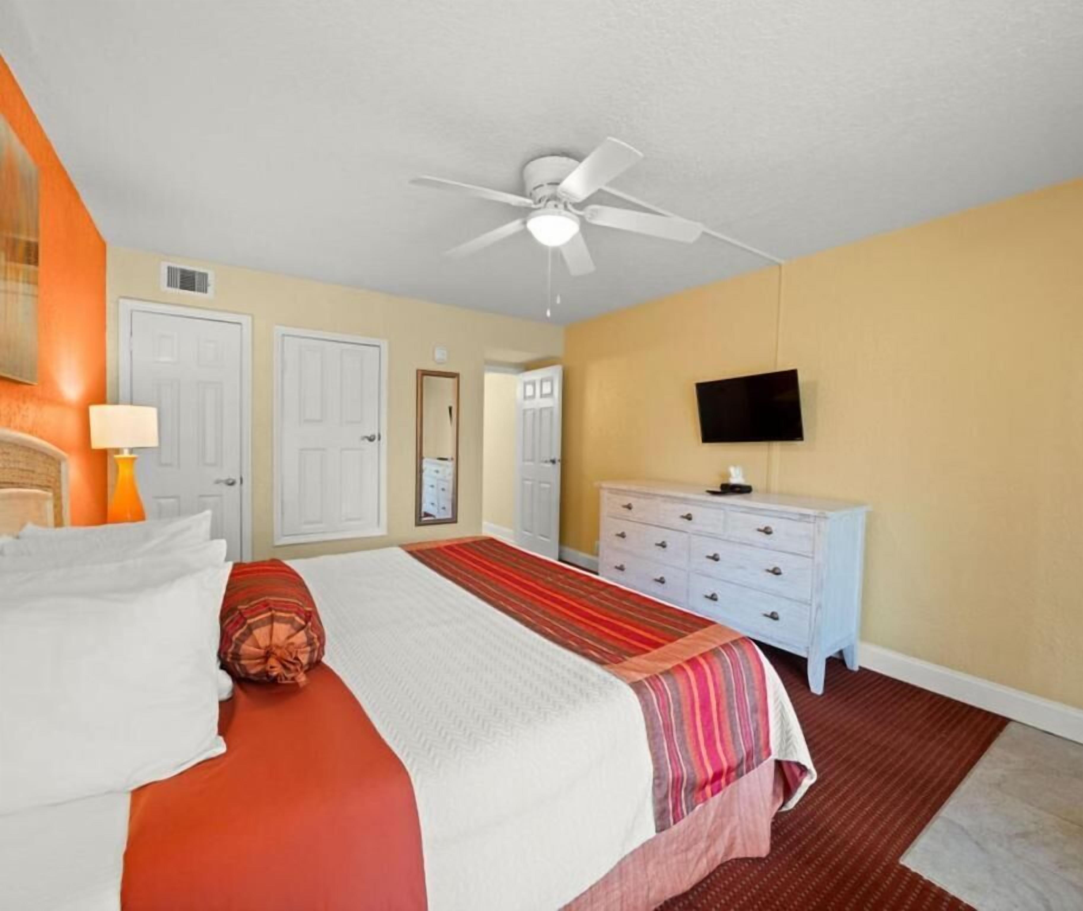Une chambre d’hôtel avec un lit, un ventilateur de plafond, une commode, un téléviseur et une porte.