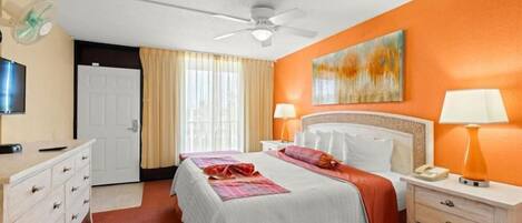 Una habitación de hotel con cama, ventilador de techo, televisor, escritorio y mesita de noche.