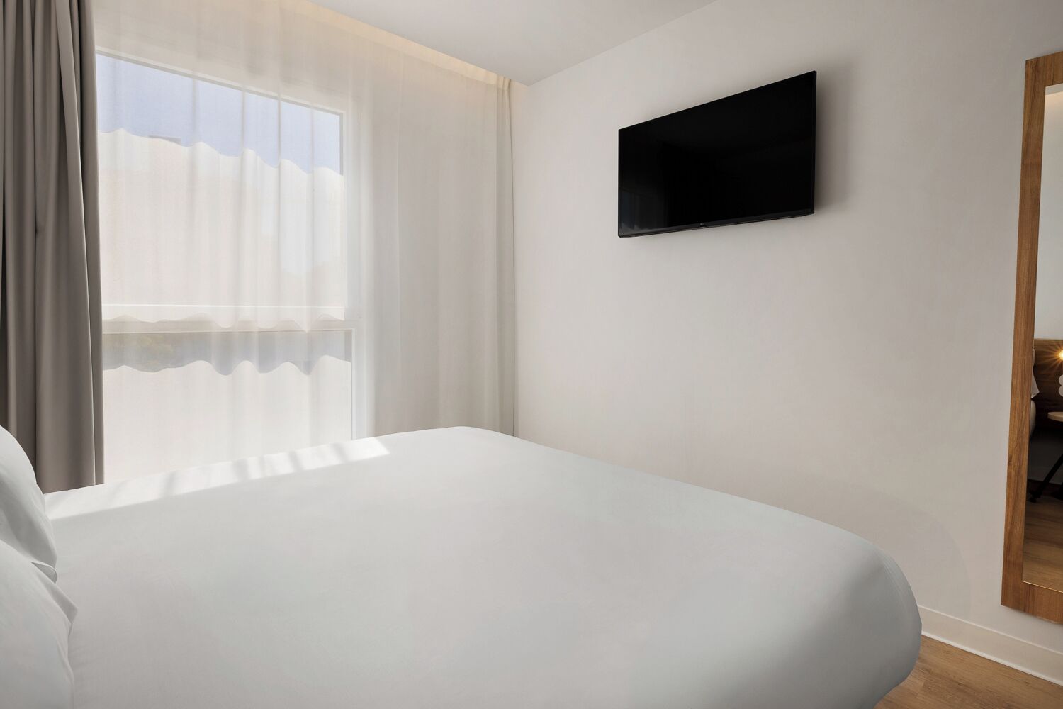 Una habitación de hotel con cama, televisión montada en la pared y ventana con cortinas.