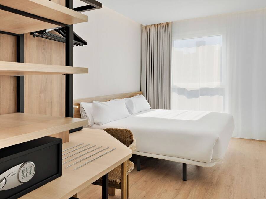 Habitación de hotel con cama, escritorio, computadora, silla y ventana con cortinas.