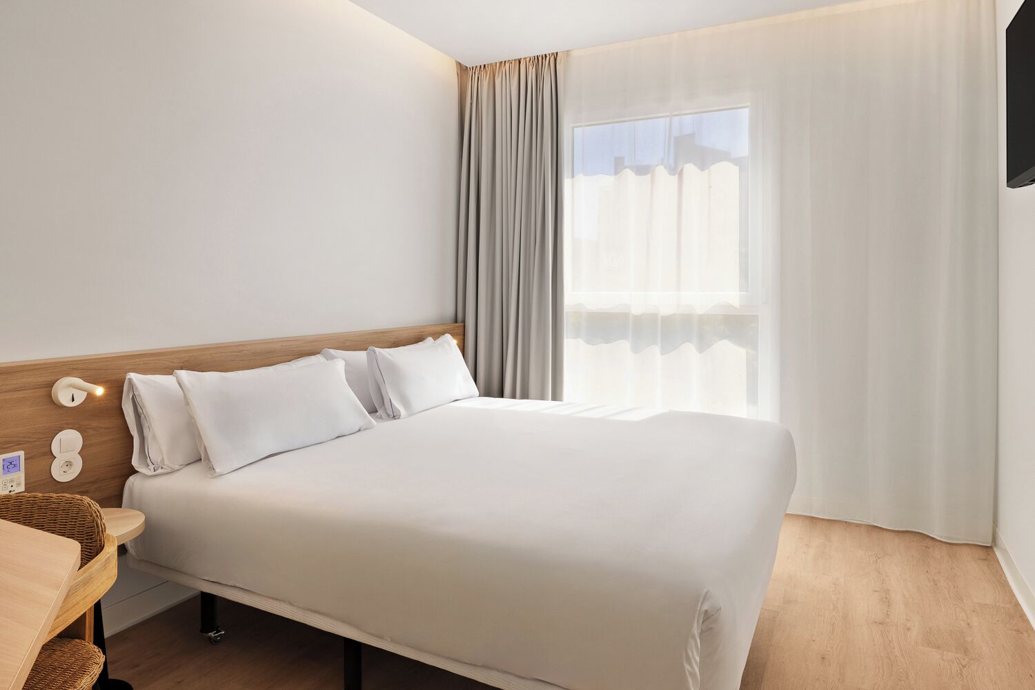 A hotel room with a large bed, a wooden headboard, a bedside table with a lamp, and a window with curtains.