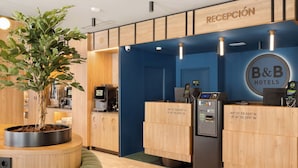Un área de recepción con un mostrador de madera, el logotipo de B&B Hotels, un quiosco de información digital y una planta en maceta.