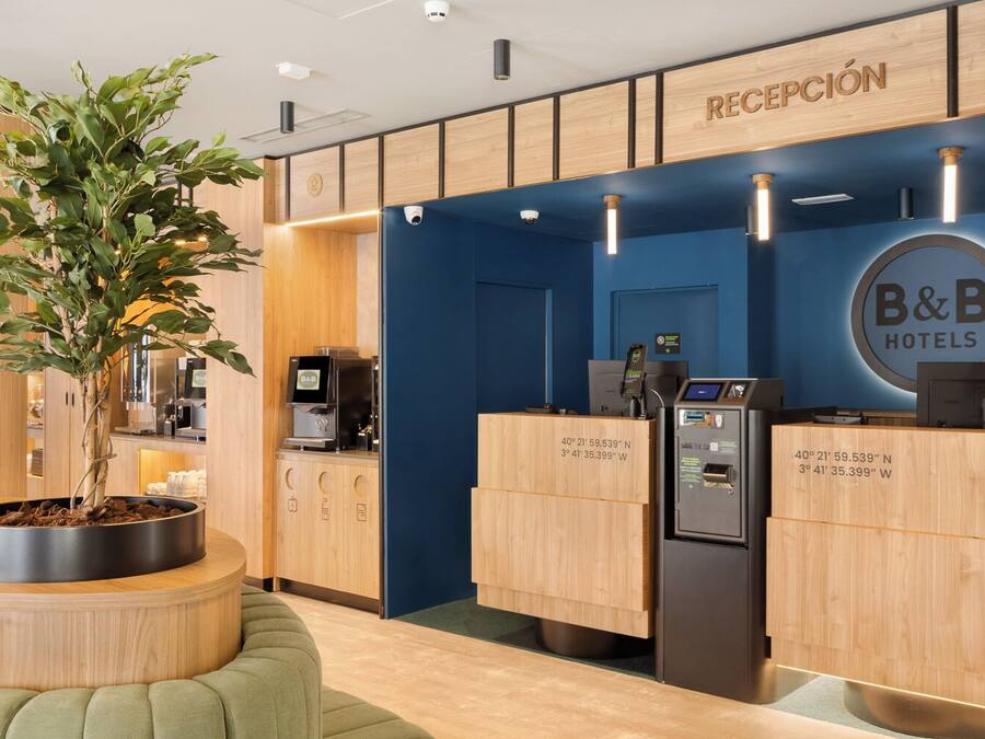 A reception area with a wooden reception desk, a B&B Hotels logo, a digital information kiosk, and a potted plant.