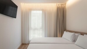 Una habitación de hotel con cama, televisor montado en la pared y ventana con cortinas.
