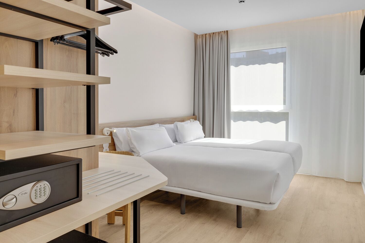 Quarto de hotel com cama, escrivaninha com computador, televisão e janela com cortinas.