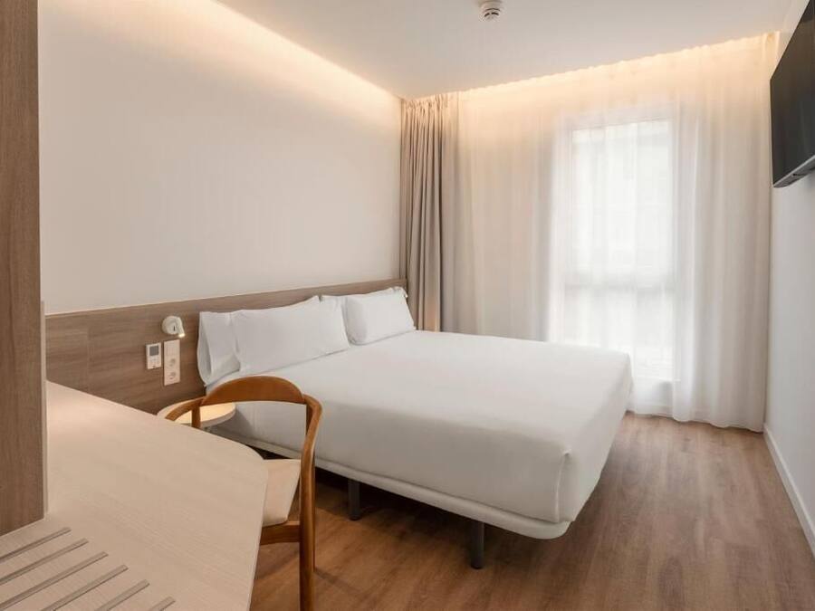 Habitación de hotel con cama, una silla, televisor montado en la pared y ventana con cortinas.