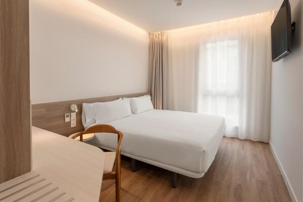 A hotel room with a bed, a chair, a TV mounted on the wall, and a window with curtains. - B&B HOTEL Madrid San Fermin (Madrid)