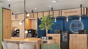 A reception area with a B&B Hotels sign, a coffee machine, seating, and a plant. - B&B HOTEL Madrid San Fermin (Madrid)