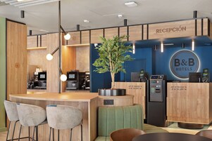 A reception area with a B&B Hotels sign, a coffee machine, seating, and a plant.