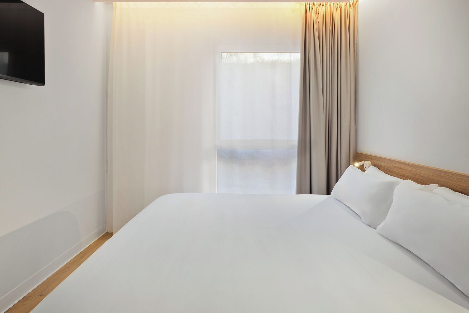 Ein Hotelzimmer mit einem großen Bett, einem Holz-Kopfteil, einem an der Wand befestigten Flachbildfernseher und einem Fenster mit Vorhängen.