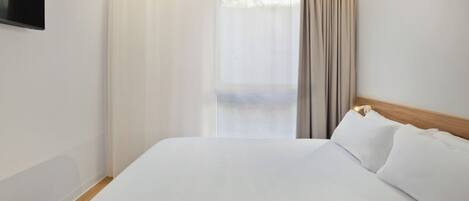 A hotel room with a large bed, a wooden headboard, a flat-screen TV mounted on the wall, and a window with curtains.