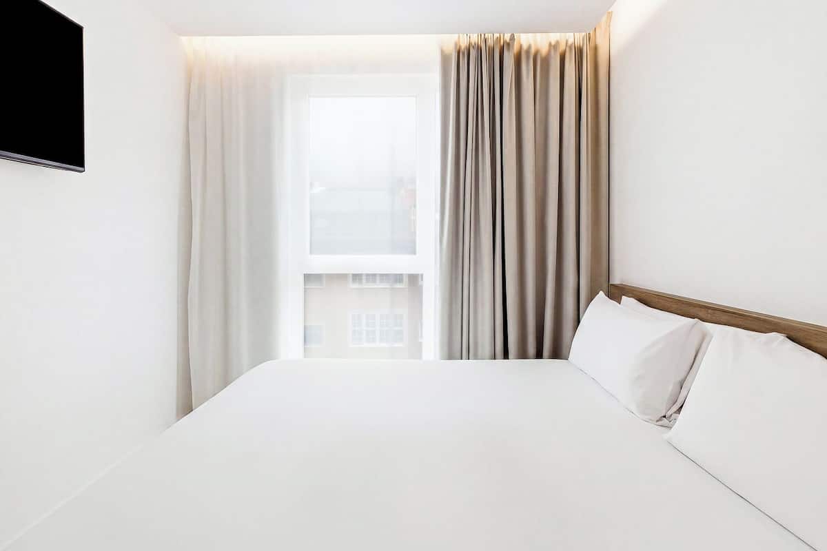 A hotel room with a bed, curtains, a TV, and a window with a view of buildings.