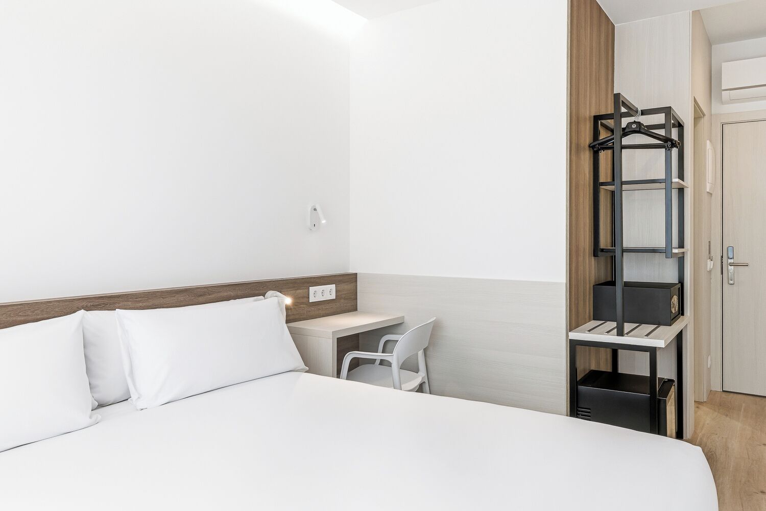 A hotel room with a bed, a desk, a chair, a wardrobe, and a hanging shelf.