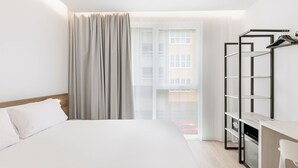Habitación de hotel con cama, escritorio y silla, perchero y vistas a edificios a través del ventanal.