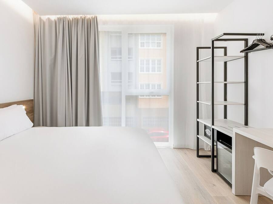 Habitación de hotel con cama, escritorio y silla, perchero y vistas a edificios a través del ventanal.