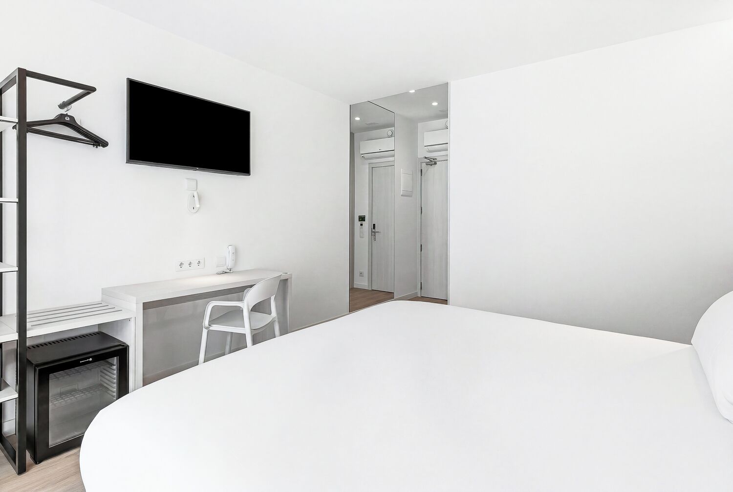 A modern hotel room with a bed, a desk with a chair, a TV mounted on the wall, and a wardrobe.