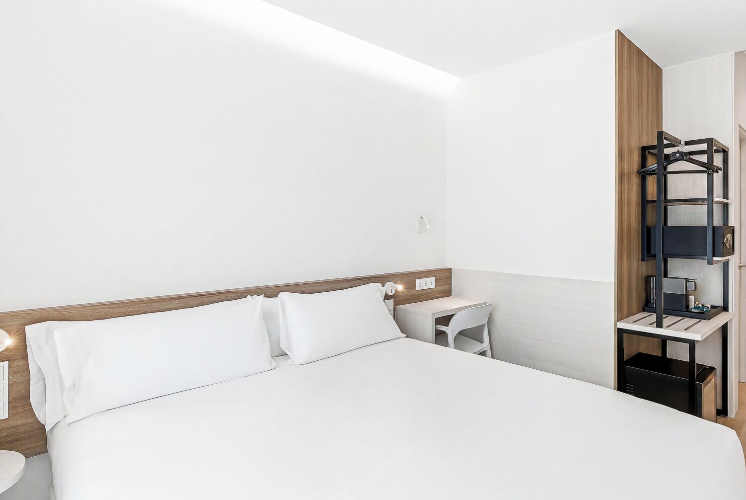 Habitación de hotel con cama, mesita de noche, silla y un estante empotrado con perchero.