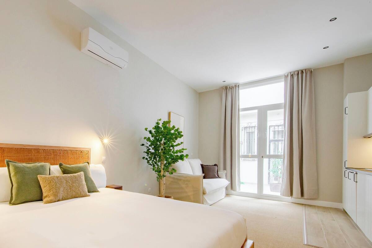 Una habitación de hotel con cama, mesitas de noche, una planta, ventana con cortinas y aire acondicionado instalado en la pared.