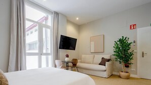 Habitación de hotel con cama, sofá, televisión, una mesita, una silla, planta en maceta y puerta con detector de incendios.