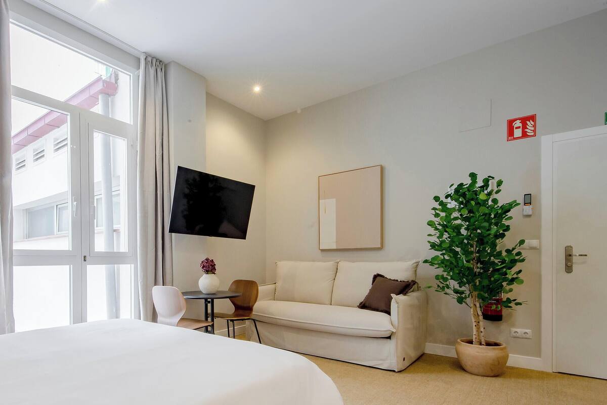 Habitación de hotel con cama, sofá, televisión, una mesita, una silla, planta en maceta y puerta con detector de incendios.