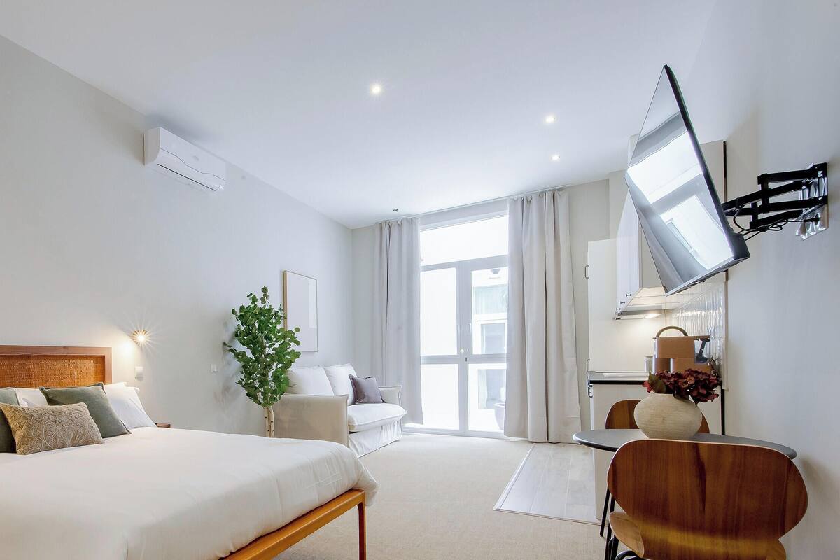 Una habitación de hotel con cama, mesita de noche, escritorio con silla, planta en maceta y un ventanal con cortinas.