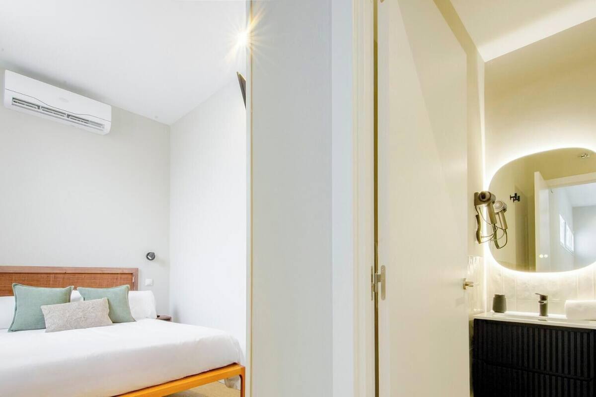 Habitación de hotel con cama, mesita de noche, aire acondicionado instalado en la pared, y baño con lavamanos, espejo y toallero.