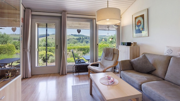 Ein modernes Wohnzimmer mit Sofa, Sessel und Tisch, großen Fenstern mit Vorhängen und Blick auf Grünflächen.