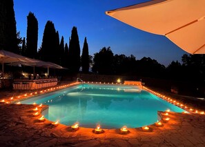 Ein Schwimmbecken, umgeben von Steinstufen, Kerzen, Sitzgelegenheiten im Freien und hohen Bäumen im Hintergrund.