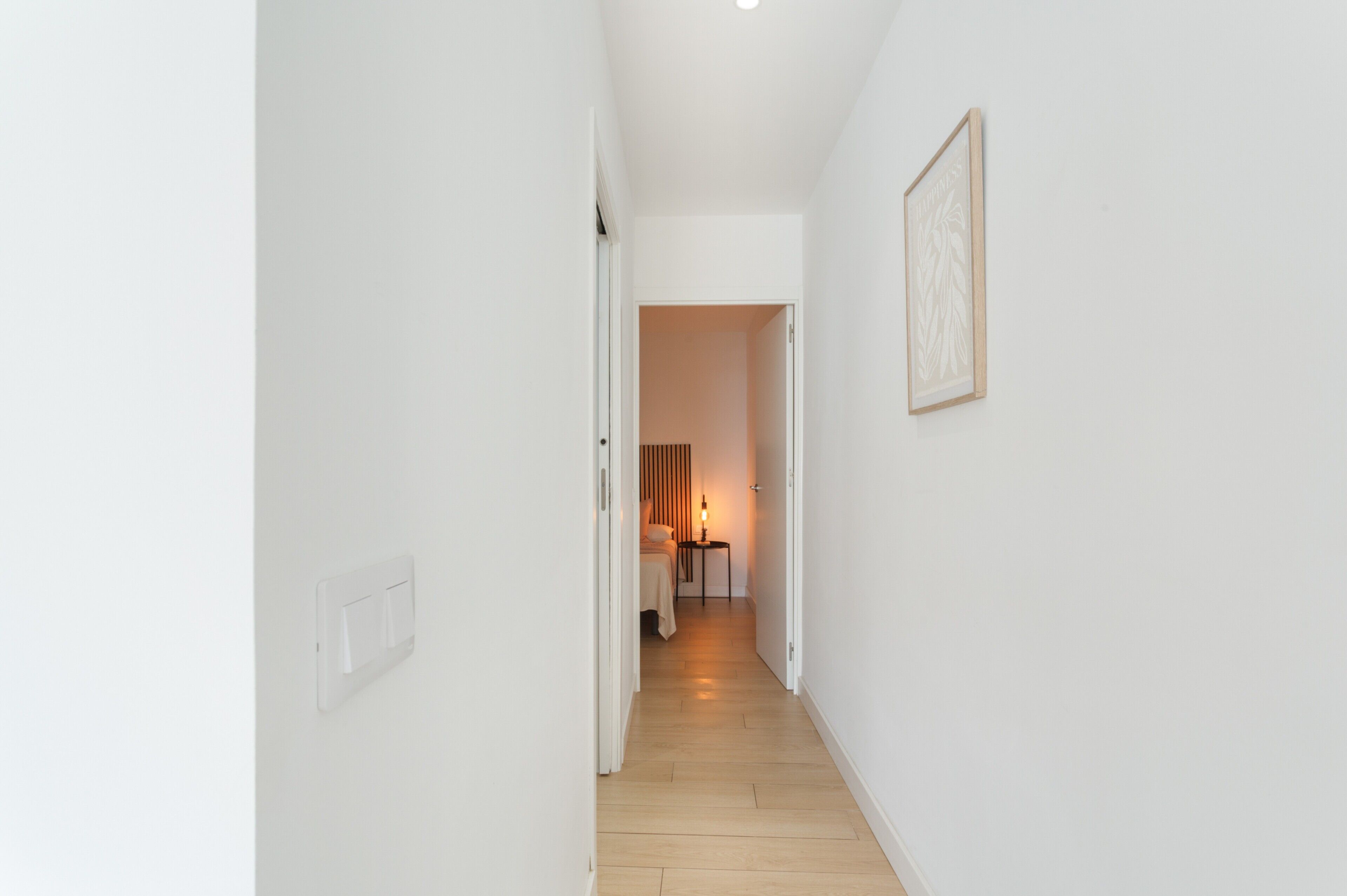 Ein schmaler Flur mit weißen Wänden, Holzboden, einem Bett mit Nachttisch und Lampe sowie einem gerahmten Bild an der Wand.