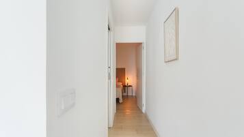 Ein schmaler Flur mit weißen Wänden, Holzboden, einem Bett mit Nachttisch und Lampe sowie einem gerahmten Bild an der Wand.