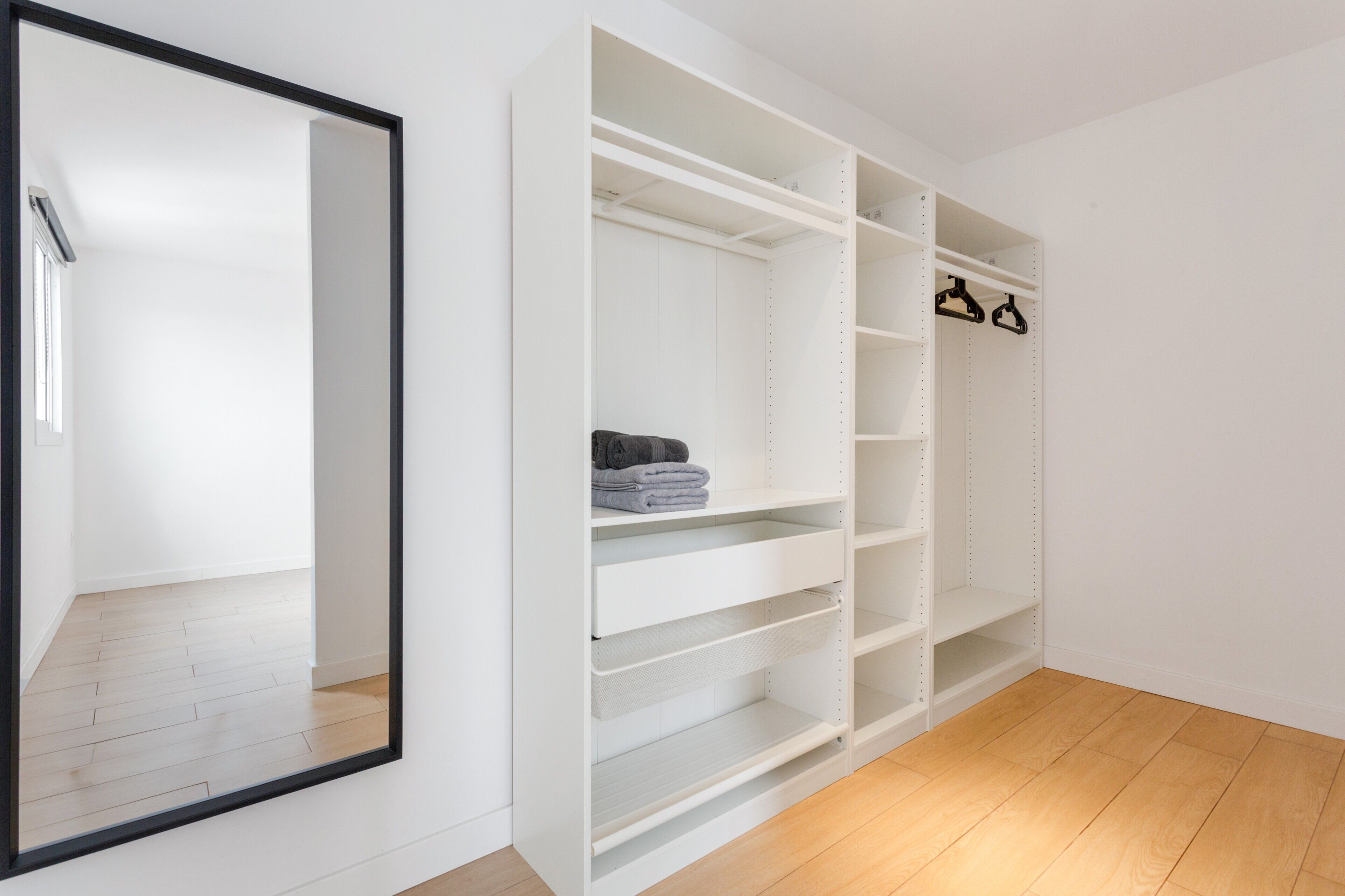 A white closet with shelves and hanging space, a mirror, and a wooden floor.