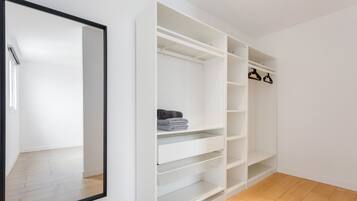A white closet with shelves and hanging space, a mirror, and a wooden floor.
