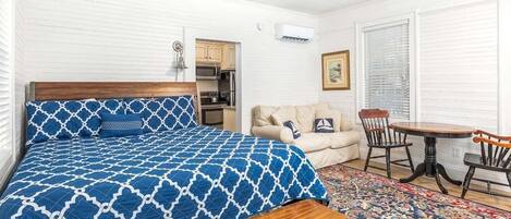 A bedroom with a bed, a wooden bench, a round table, a sofa, and a kitchen area in the background.