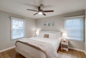 Ein Schlafzimmer mit einem Bett, einem Deckenventilator, zwei Nachttischen und einem Fenster mit Jalousien.