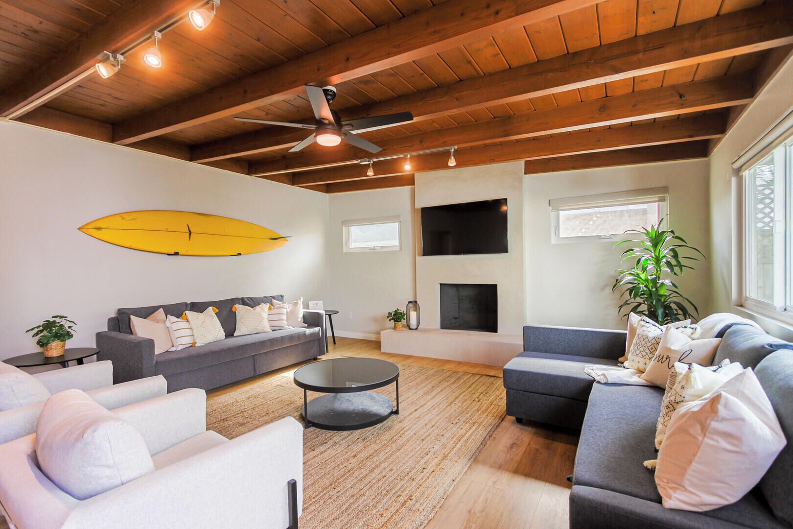 A living room with a sofa, armchairs, a coffee table, a fireplace, a TV, and a surfboard on the wall.