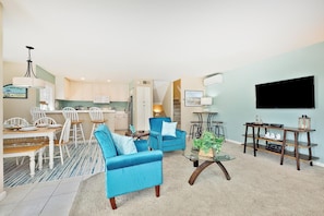 Ein Wohnzimmer mit blauem Sofa, Couchtisch, Fernseher und Küchenzeile mit weißen Schränken.