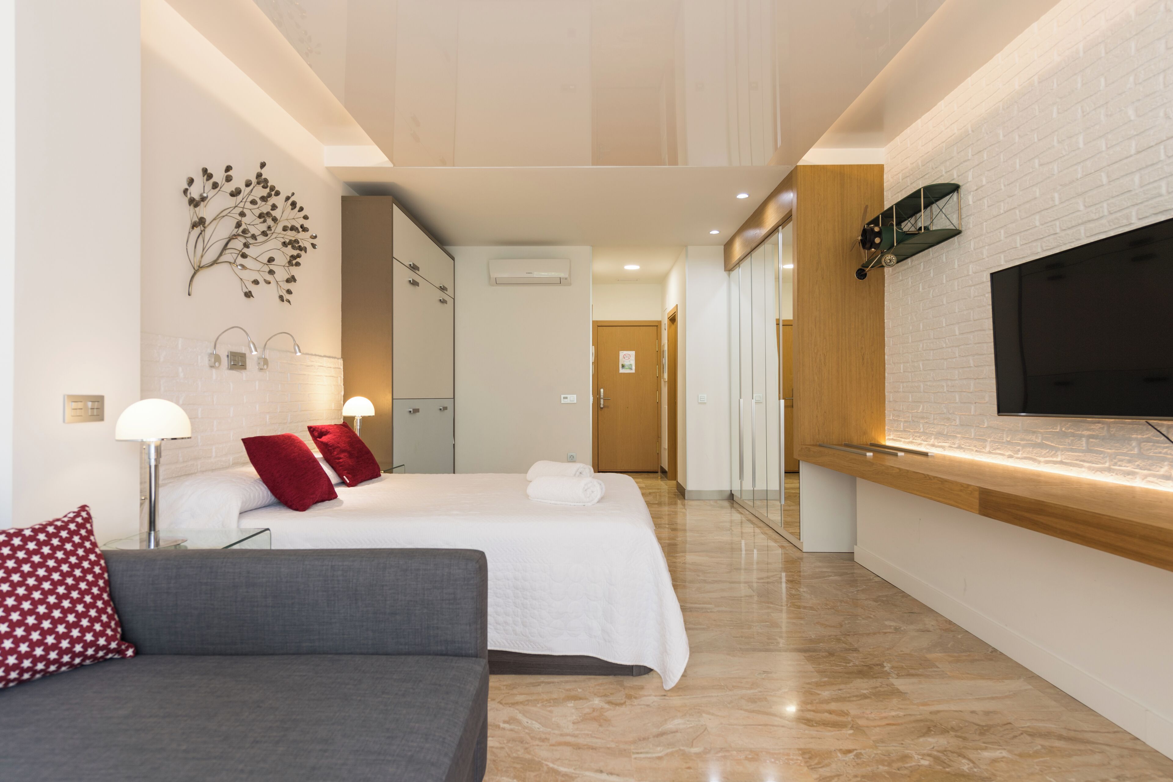 A modern hotel room with a bed, a sofa, a TV, and a bathroom visible through an open door.