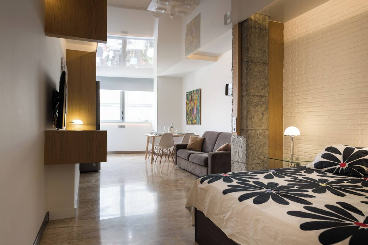 A modern hotel room with a bed, a desk, a sofa, and a dining area.