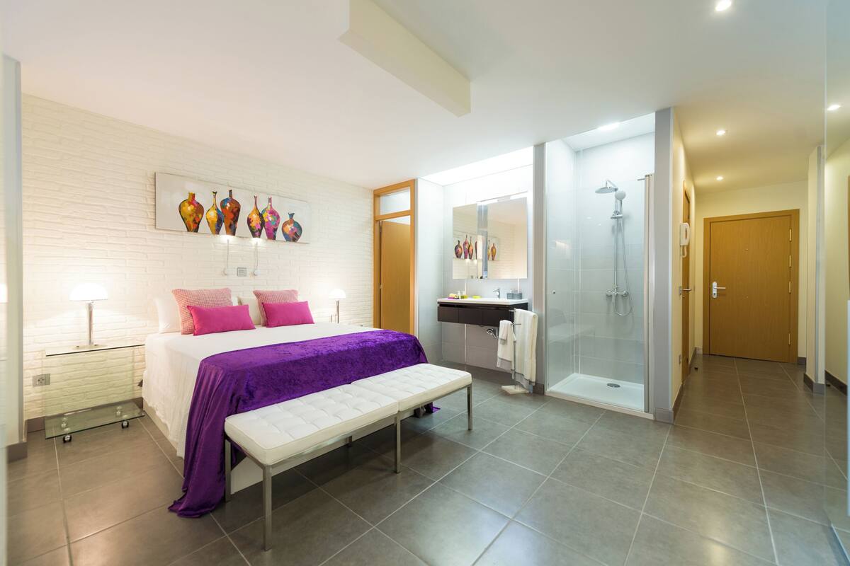 A modern bedroom with a bed, bench, bedside tables, a shower, and a bathroom with a sink and mirror.