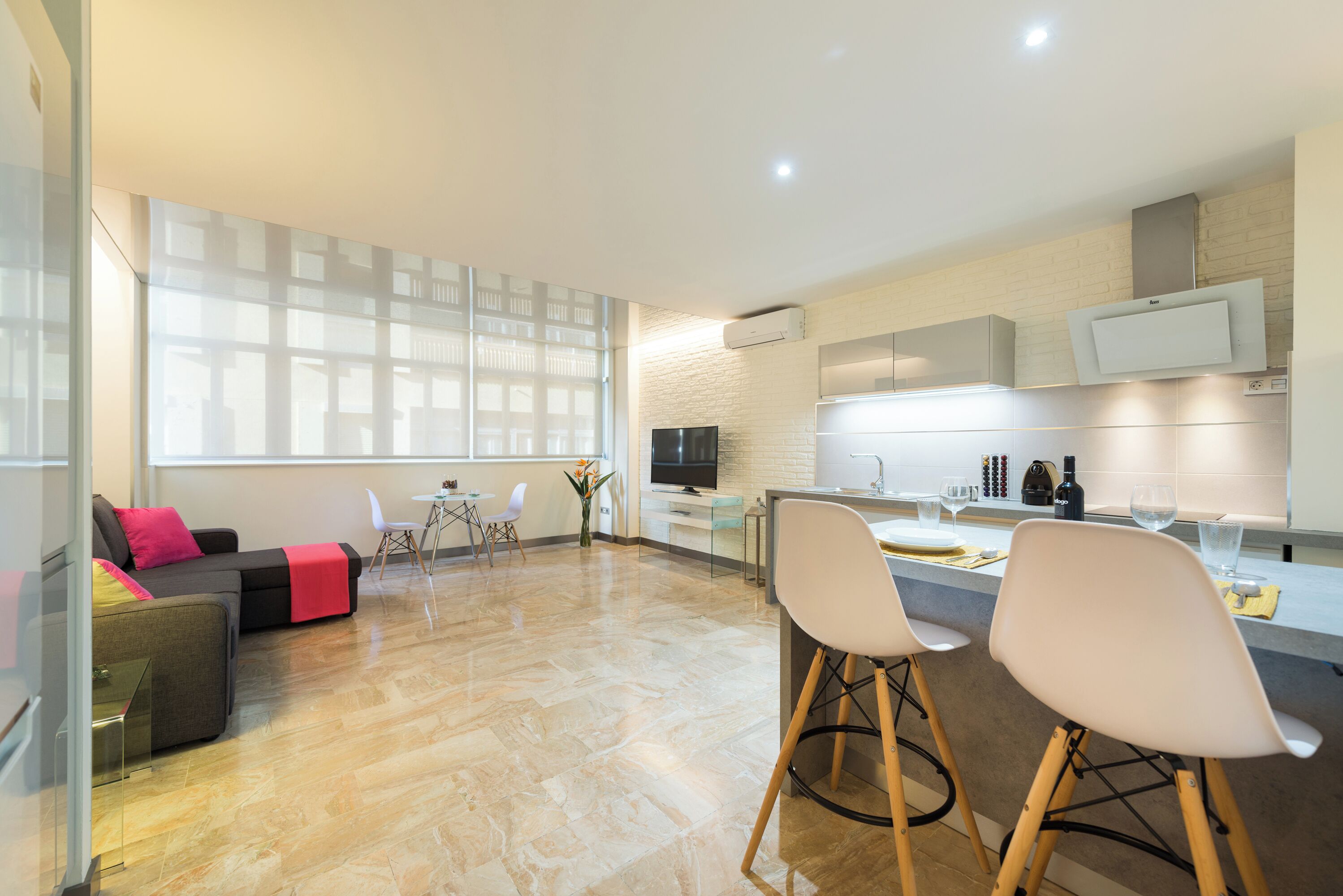 A modern kitchen with a flat-screen TV, a dining area with bar stools, and a sofa with a red cushion.