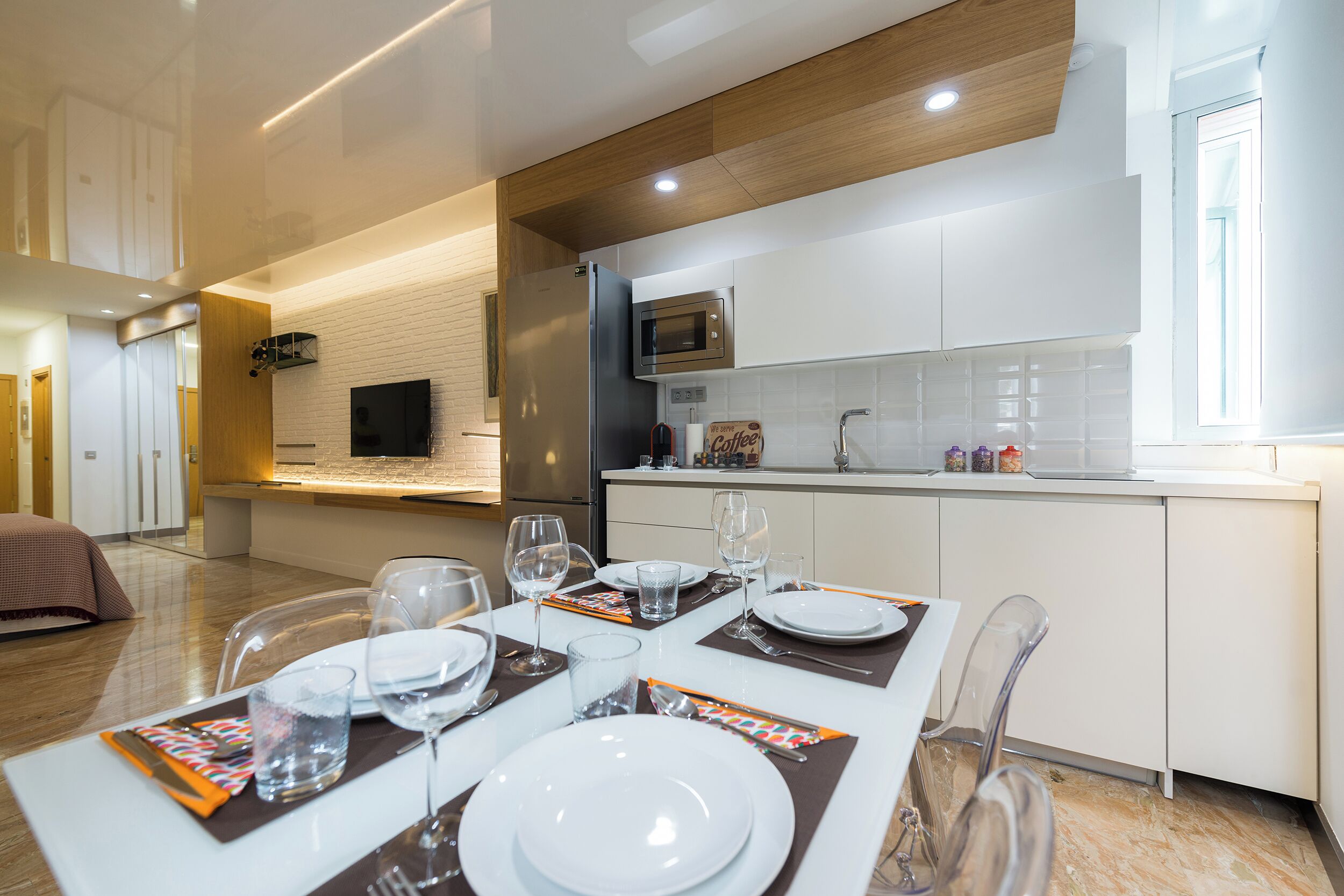 A modern kitchen with white cabinets, a built-in microwave, and a dining area set for four with clear chairs and a white table.