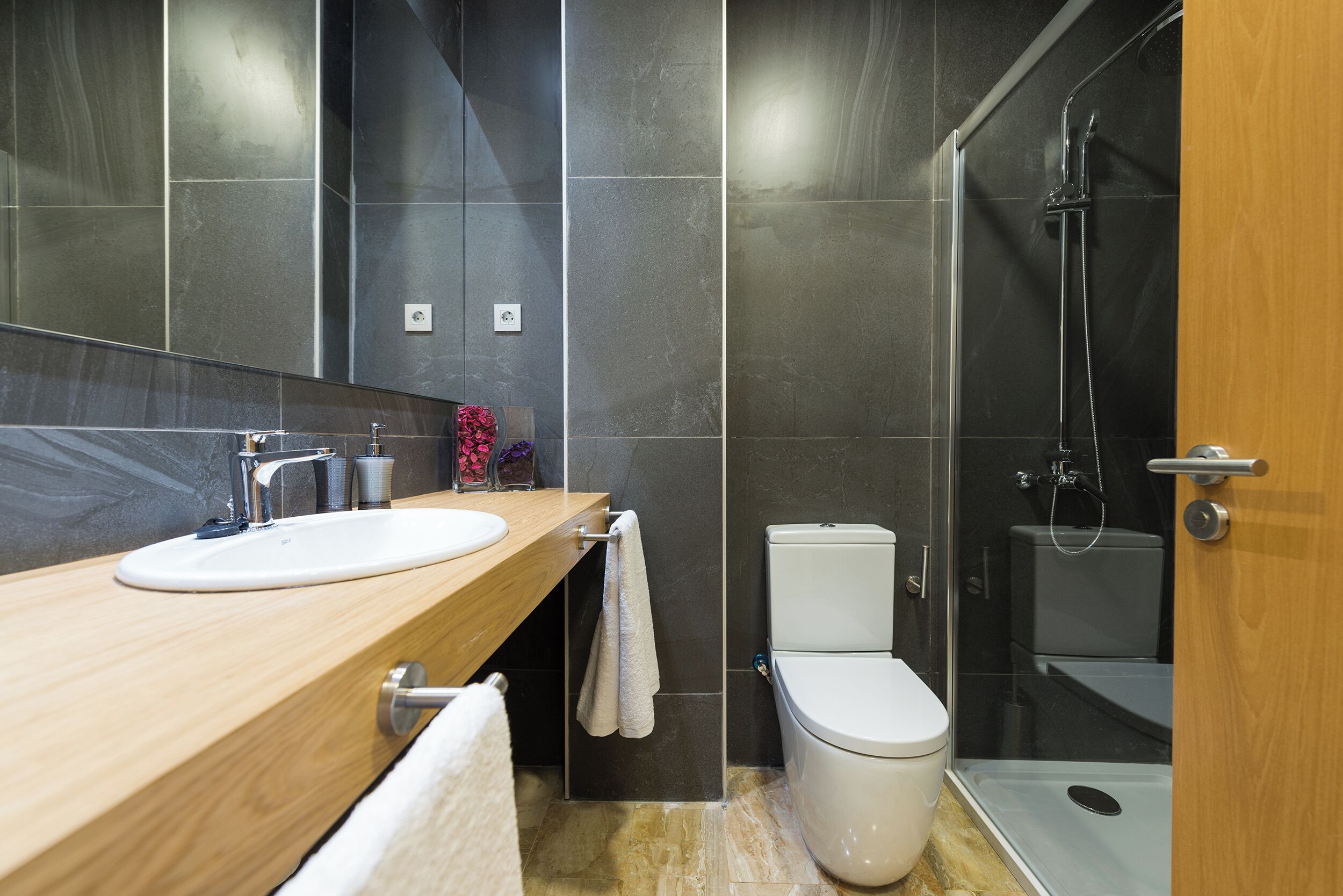Un baño moderno con tocador de madera, lavamanos blanco, inodoro y ducha con cerramiento de vidrio.