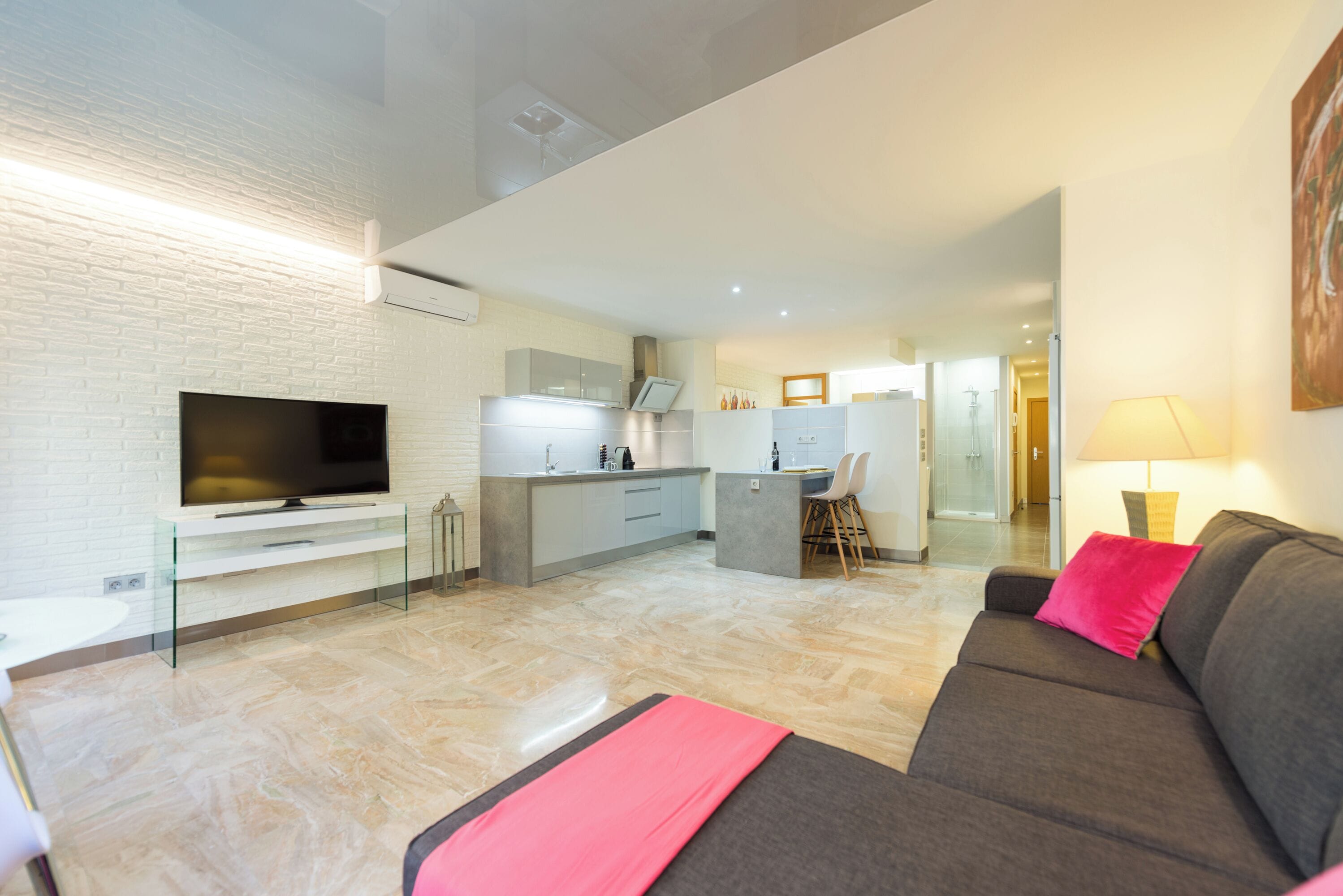 A modern living room with a flat-screen TV, a grey sofa with a pink pillow, a glass coffee table, and a kitchen area in the background.