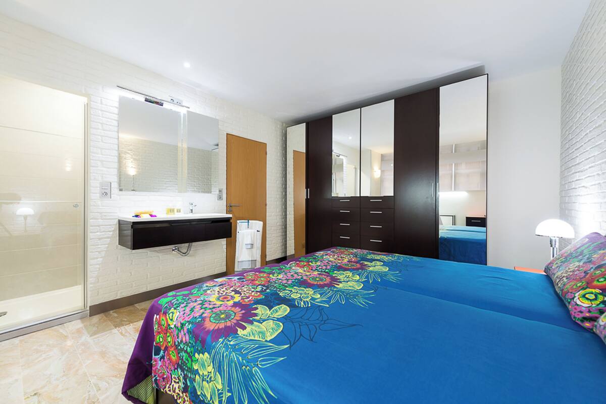 A bedroom with a large bed, a bathroom with a glass shower, a wardrobe, and a mirror.