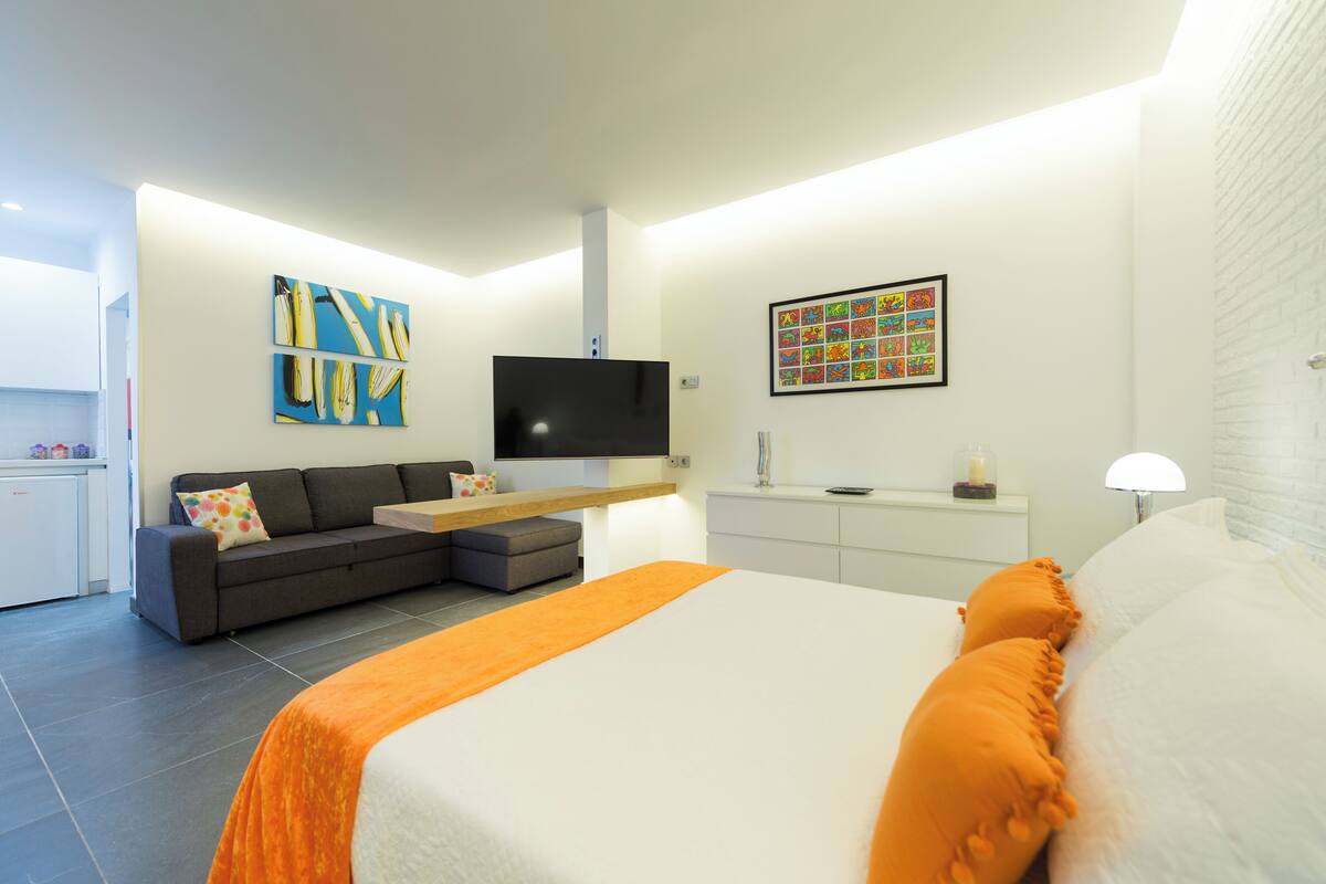 A modern hotel room with a bed, sofa, TV, artwork, and a small kitchenette.