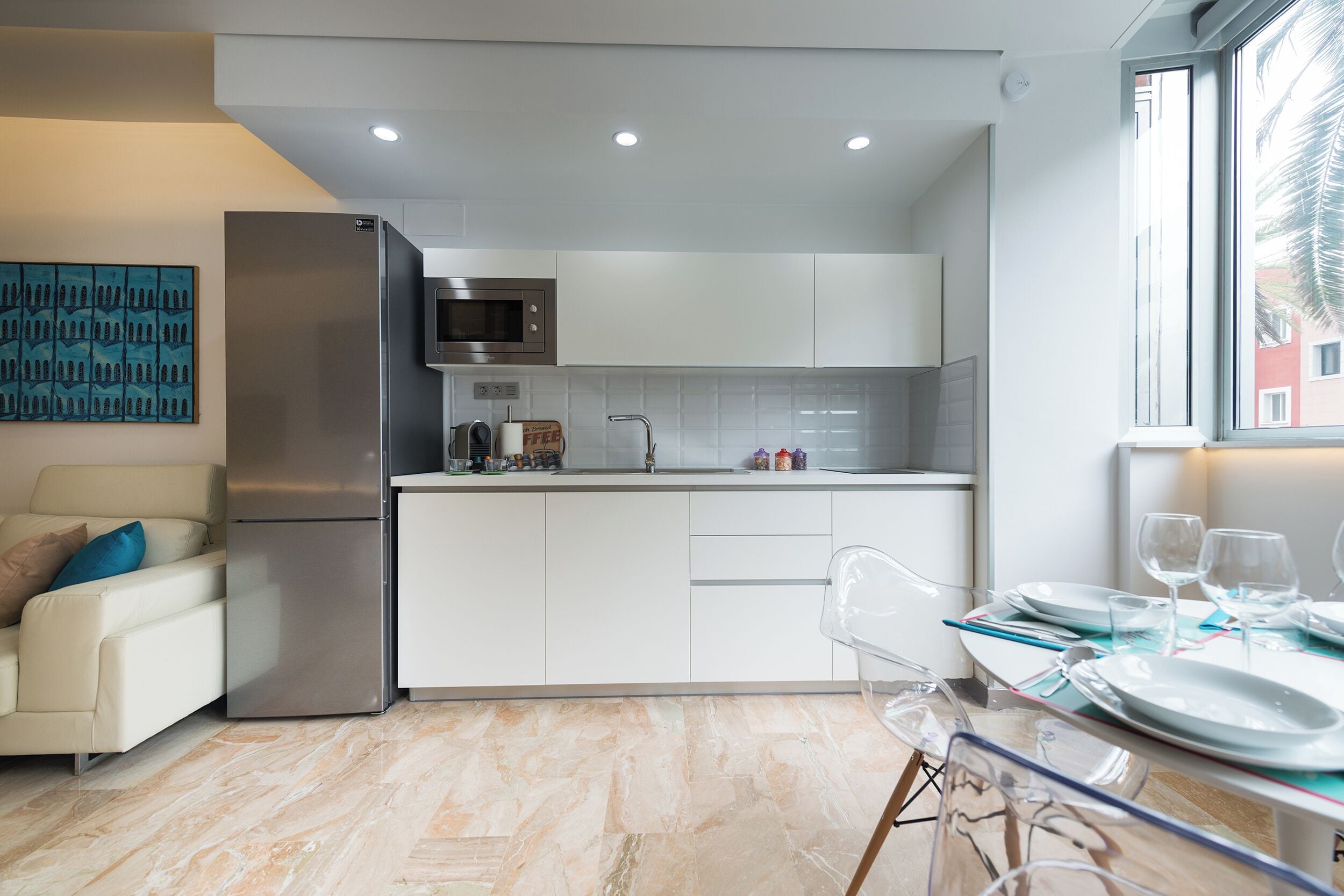 Una cocina moderna con refrigerador, microondas y gabinetes blancos. Hay un comedor con una mesa lista para dos personas y una ventana con vistas.
