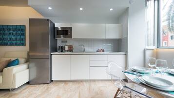 Una cocina moderna con refrigerador, microondas y gabinetes blancos. Hay un comedor con una mesa lista para dos personas y una ventana con vistas.