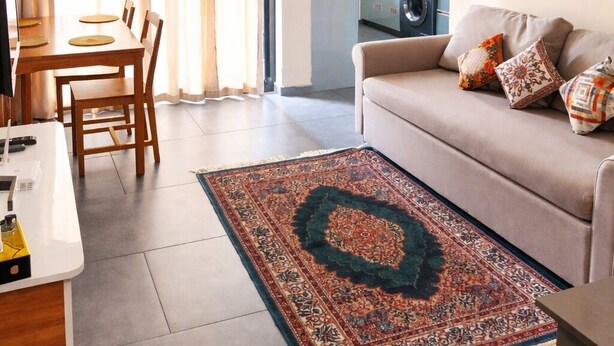 A living room with a sofa, a dining table, a kitchen in the background, a ceiling fan, and a patterned rug.