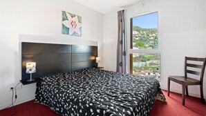 A bedroom with a large bed, a headboard, bedside lamps, a chair, a window with a view, and a painting on the wall. - Flat with a balcony offering views of the mountains and the swimming pool (Grasse)