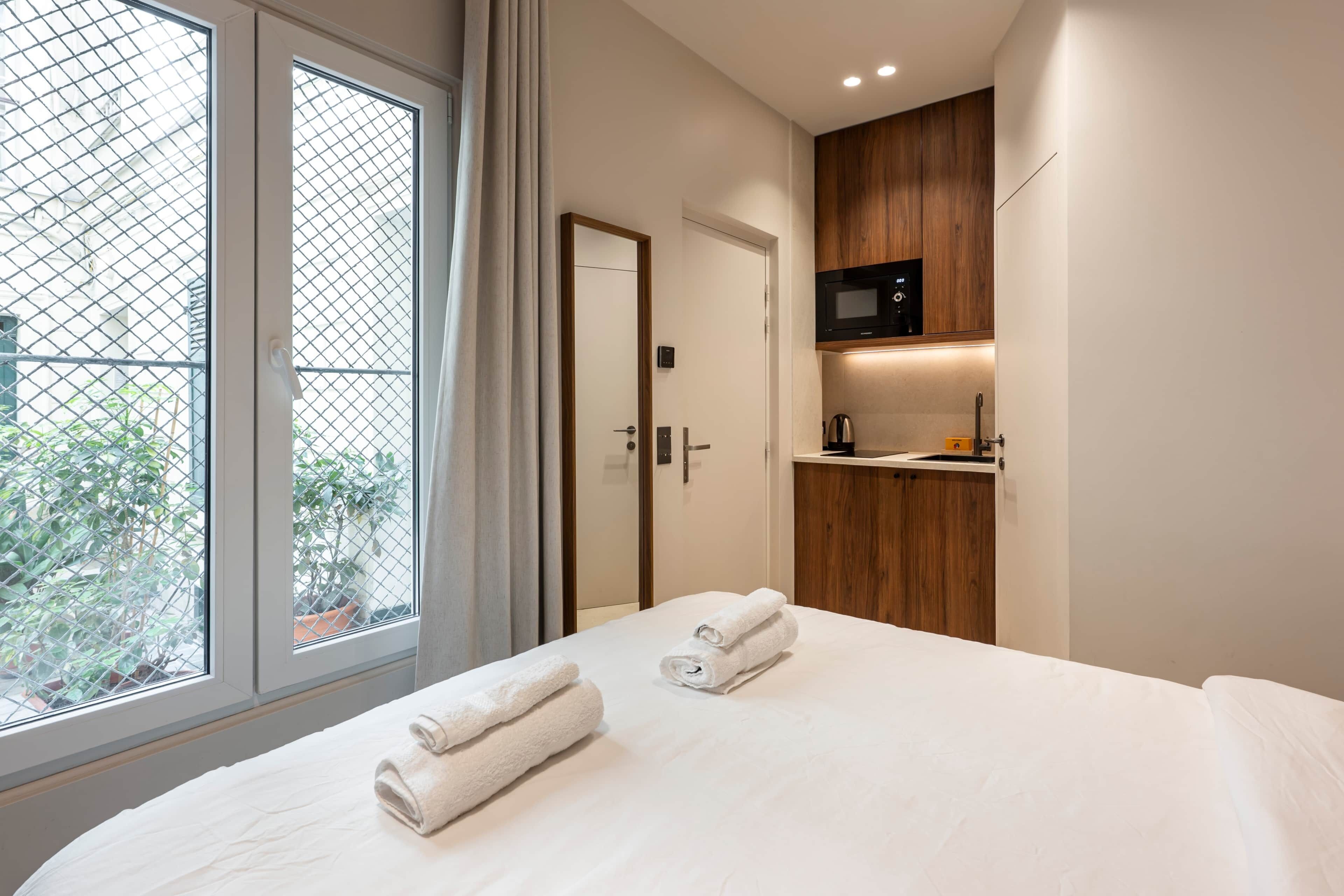 A hotel room with a bed, two towels, a window with a view, a door, and a kitchenette area.
